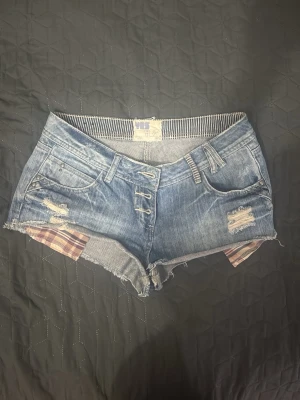 yes yes låg midja Blå jeansshorts med slitningar - Säljer ett par låg midjade blå yes yes jeansshorts med slitna detaljer och råa kanter. Shortsen har knappar framtill, bälteshällor och fickor både fram och bak. Insidan av benen har ett rutigt tyg som sticker fram för en cool kontrast.