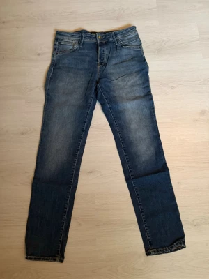 Blå raka jeans med fem fickor från Jack&Jones - Säljer ett par klassiska blå jeans med rak passform och fem fickor. Jeansen har en snygg tvätt med lätt slitning framtill och bak, samt en brun patch vid linningen bak. Tillverkade i denim med dragkedja och knapp framtill.