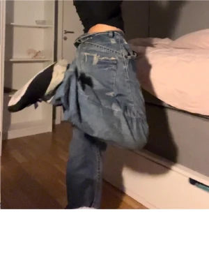 Baggy blå jeans med tryck bak - Säljer ett par baggy blå jeans med slitningar och ett stort svart tryck på ena bakfickan. Jeansen har en lös passform och klassisk femficksdesign. Perfekta för en avslappnad och trendig streetwear-look.