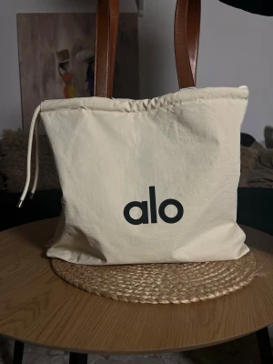 Alo yoga tote bag Beige tygväska med bruna handtag - Snygg beige handväska från alo med bruna handtag i skinnimitation och praktisk snörning upptill. Väskan är rymlig och har en enkel, stilren design med alo-logga framtill. Perfekt för dig som gillar minimalistisk stil.