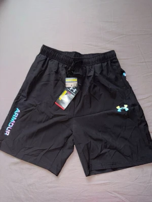 Svarta reflektiva träningsshorts från Under Armour - Svarta träningsshorts från Under Armour med elastisk midja och snörning. Logga i vitt och blått på ena benet samt reflekterande detaljer bak. Perfekta för sport eller gym. Lätta och bekväma i syntetmaterial.