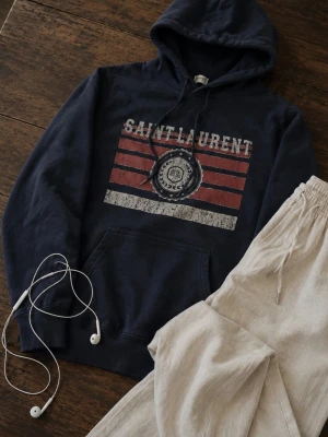 Mörkblå hoodie från Saint Laurent - Säljer en mörkblå hoodie från Saint Laurent med tryck framtill i vitt och rött samt texten 'SAINT LAURENT'. Hoodien har huva med dragsko och en stor magficka. Materialet är mjukt och skönt, perfekt för en avslappnad stil. Passar S.