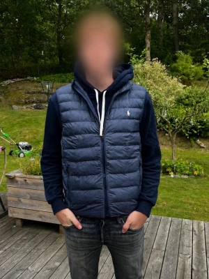 Marinblå dunväst från Polo Ralph Lauren - Snygg mrinblå dunväst från Polo Ralph Lauren med klassisk quiltad design, hög krage och dragkedja framtill. Västen har två fickor med dragkedja och den ikoniska vita logon på bröstet. Perfekt för lager-på-lager och streetstyle.