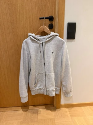 Ljusgrå zip hoodie från Ralph Lauren - Ljusgrå hoodie med dragkedja från Ralph Lauren. I fint skicka, dvs 8/10. Inga defekter. Sparsamt använd. Storleken är M