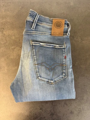 Ljusblå Replay Anbass, stl 29/34 - Säljer ett par ljusblå jeans med raka ben och snygga slitningar. Jeansen har klassisk femficksdesign, kontrastsömmar och en patch i läderimitation bak i midjan. Materialet är denim i bomull med en skön tvättad look.
