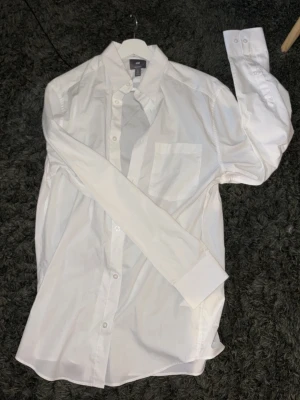 Klassisk vit skjorta med lång ärm - Säljer en stilren vit skjorta med lång ärm och bröstficka. Skjortan har knappar framtill och klassisk krage, perfekt för en clean och enkel look. Tillverkad i bomull för en bekväm känsla.