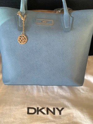 Rea! Fantastiskt fin och oanvänd DKNY handväska – himmelsblå – 100 % äkta skinn - Sommarfynd! Originalpris 2.795 kr.  Oanvänd tote bag, perfekt storlek för jobb eller studier, från DKNY i en superfin ton av himmelsblått. 100 % äkta högkvalitativt skinn.  Ett rymligt huvudfack med dragkedja. Inuti: Ett mindre fack med dragkedja + två öppna fack. Dubbla bärhandtag, en avtagbar axelrem.  Eleganta detaljer i guldfärgad metall, inklusive en DKNY-logotyp och en rund tillhörande berlock / bag charm med "DKNY Donna Karan New York" präglat.  Otroligt bra pris, passa på! Dustbag ingår.