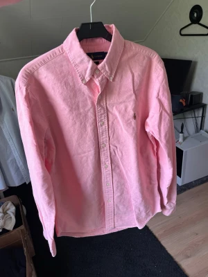 Rosa skjorta med knappar - Säljer en klassisk rosa skjorta med knappar framtill och button-down krage. Skjortan har lång ärm och en liten broderad logga på bröstet. Perfekt för dig som vill ha en stilren och enkel look.