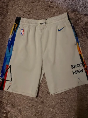 Brooklyn Nets vita basketshorts - Säljer ett par vita basketshorts med färgglada grafiska detaljer längs sidorna och broderad text 'BROOKLYN NEW'. Shorts med elastisk midja, blå Nike-logga och NBA-märke. Perfekta för dig som gillar streetwear och sportig stil.