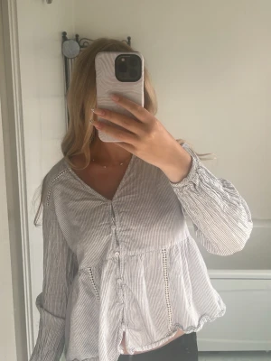 Randig blus från ONLY med volang - Säljer en ljusblå och vit randig blus från ONLY med knappar framtill och volang nedtill. Blusen har v-ringning, långa ärmar med resår vid ärmslut och fina spetsdetaljer vid axlar och midja. Perfekt till jeans eller kjol för en fräsch look.