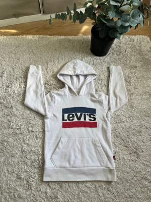 levi's hoodie - Säljer en vit hoodie från Levi's med stor logga i blått, vitt och rött på bröstet. Hoodien har huva, magficka och ribbade muddar. Perfekt för en chill och avslappnad stil. Materialet är mjukt och skönt, troligen bomullsmix. Några frågor? Skriv till mig, svarar så snabbt jag kan!🙌8 år gammal strl