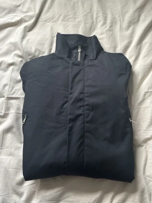 Svart jacka från Elvine med dragkedja - Snygg svart jacka från Elvine med hög krage och dragkedja framtill. Jackan har två sidofickor med dragkedja och ett stilrent, minimalistiskt utseende. Perfekt för dig som gillar enkel och clean design.