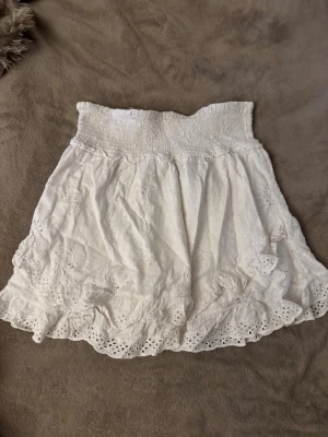 Vit broderad volangkjol - Söt vit kjol med bred smock i midjan och broderade detaljer. Kjolen har volanger och spetskant längst ner vilket ger en romantisk och bohemisk känsla. Perfekt för sommardagar och festivaler.