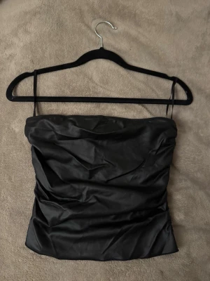 Svart tubtopp - Snygg svart tubtopp i glansigt skinliknande material med smala axelband. Toppen har en croppad passform och är perfekt för att skapa en trendig look. Enkel och stilren design som passar till många olika outfits.