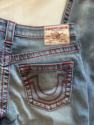 True Religion ljusblå jeans med röd söm - Ljusblå jeans från True Religion med ikoniska bakfickor och tydliga röda kontrastsömmar. Klassisk femficksmodell och bootcut-ben. Snygga detaljer och patch bak i midjan. Perfekta för dig som gillar street och statement-jeans. Modellen heter BECCA mid rise bootcut 🌟storleken är W27, innerbenslängden är 83cm 🔥🔥 jeansen är nyskick men kommer tyvvär utan prislapp