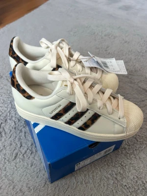Adidas Superstar med leoparddetaljer - Säljer ett par Adidas Superstar sneakers i vitt med ikoniska tre ränder och häl i leopardmönstrad mocka. Klassisk snäcktå och vita skosnören. Skorna är i lågt utförande och har en fräsch och trendig look som sticker ut.