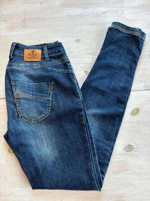 Blå jeans från Pulz Jeans - Snygga blå jeans från Pulz Jeans med klassisk femficksdesign och dekorativa nitar på bakfickan. Jeansen har en smal passform och är tillverkade i slitstarkt denim med snygga slitningar och kontrastsömmar. Perfekta till en avslappnad vardagsstil.