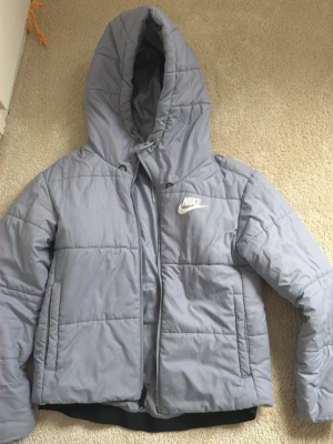 Ljusblå pufferjacka från Nike - Snygg ljusblå pufferjacka från Nike med huva och dragkedja framtill. 