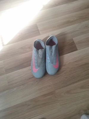 Gråa Nike fotbollsskor med rosa swoosh - Säljer ett par gråa Nike fotbollsskor med en stor rosa swoosh på sidan och perforerade detaljer upptill. Skorna har låg profil och dobbar för bra grepp på planen. Snygg design som sticker ut på matchen.