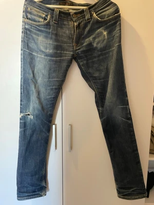 Blå jeans från Nudie Jeans - Snygga blå jeans från Nudie Jeans med slitningar och en liten reva på vänster ben. Klassisk femficksmodell med orangea sömmar och broderade bakfickor. Jeansen har normal passform och är tillverkade i bomull.