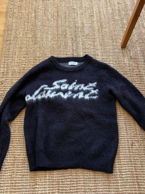 Svart Saint Laurent stickad tröja - Säljer en svart stickad tröja från Saint Laurent med vit grafisk text på bröstet. Tröjan har rund halsringning och är gjord i ett fluffigt, mjukt material som ger en lyxig känsla. Perfekt för dig som vill ha något unikt och trendigt i garderoben.