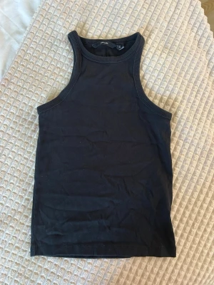 Svart ribbad linnetopp - Snygg svart ribbad linnetopp med hög hals och ärmlös design. Perfekt att styla till jeans eller kjol för en clean och enkel look. Materialet känns mjukt och stretchigt, vilket gör toppen bekväm att bära hela dagen.