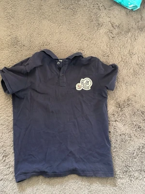 Mörkblå Moncler pikétröja - Snygg mörkblå pikétröja från Moncler med klassisk krage och två knappar framtill. Tröjan har korta ärmar och ett tydligt Moncler-märke broderat på bröstet. Perfekt för en stilren och avslappnad look.