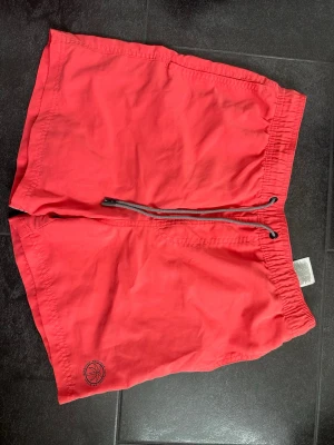 Röda badbyxor från Jack & Jones - Snygga röda badbyxor från Jack & Jones med elastisk midja och svart dragsko. Innerbyxa i mesh för extra komfort och en liten palm-brodyr på benet. Perfekta för stranden eller poolen.
