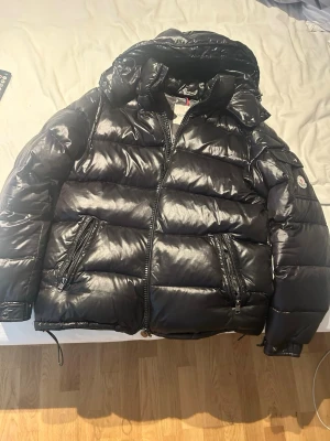 Svart glansig dunjacka med huva - Säljer en svart, glansig dunjacka från Moncler med stor huva och två dragkedjeförsedda fickor framtill. Jackan har breda kanaler och en klassisk Moncler-logga på ärmen. Perfekt för kalla vinterdagar och riktigt snygg streetstil.