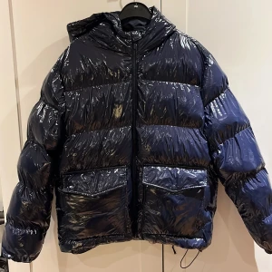 Marinblå pufferjacka från Lawrence Grey - Snygg marinblå pufferjacka från Lawrence Grey med glansig finish, huva och stora fickor framtill. Jackan har dragkedja hela vägen och en riktigt puffig look som håller dig varm under kalla dagar. Perfekt för dig som gillar streetstyle och vill sticka ut.