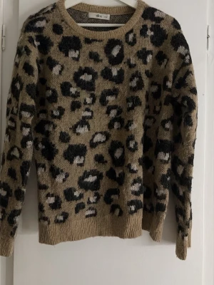 Leopardmönstrad stickad tröja - Stickad tröja från ellos med leopardmönster i beige, svart och grått. Tröjan har rund halsringning, långa ärmar och ribbade muddar vid ärmslut och nederkant. Perfekt för dig som vill ha en trendig och mysig look.