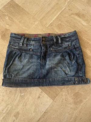 Blå jeanskjol med knappar - Snygg blå jeanskjol i kort modell med dubbla knappar framtill, bälteshällor och dekorativa fickor både fram och bak. Kjolen har slitna detaljer och knappdetaljer vid nederkanten för en cool look. .