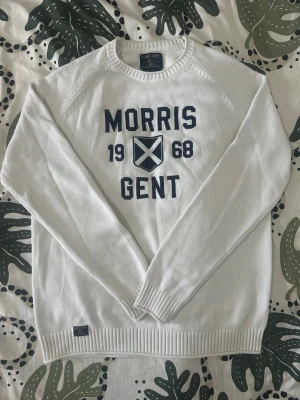 Vit stickad tröja Morris Gent - Säljer en vit stickad tröja från Morris Gent med mörkblå text och emblem på bröstet. Tröjan har rund halsringning, långa ärmar och ribbade muddar vid ärmslut och nederkant. Perfekt för dig som gillar klassisk och stilren design.