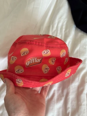 Röd bucket hat med gifflar-mönster - Cool röd bucket hat med tryck av gifflar och loggan 'gifflar' runtom hela hatten. Hatten har en klassisk form med bred brätte och är tillverkad i ett lätt tygmaterial. Perfekt för dig som vill sticka ut med en unik accessoar.