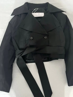 Kort svart trenchcoat från Eva Jonsson - Stilren kort svart trenchcoat från Eva Jonsson med bred krage, dubbelknäppning och knytskärp i midjan. Jackan har långa ärmar med knappdetaljer vid ärmsluten och är tillverkad i ett slätt tyg som ger en modern look.