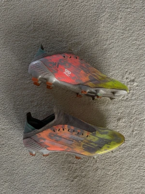 Adidas färgglada fotbollsskor - Säljer ett par Adidas fotbollsskor med låg profil och snörning. Skorna har en färgstark design i gult, orange och grått med svart mönster. Ovandelen är i mesh-liknande material och sulan har fasta dobbar i plast. Perfekta för dig som vill sticka ut på planen!
