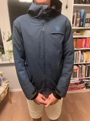 Blå vindjacka från Rossignol - Snygg blå vinterjacka från Rossignol med huva och dragkedja framtill. Jackan har en bröstficka med logga, justerbara muddar med kardborre och stort Rossignol-tryck på ena ärmen. Perfekt för blåsiga och kalla vinterdagar och outdoor-aktiviteter.