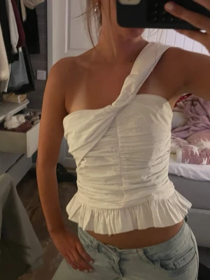 Vit oneshoulder topp med volang - Säljer en vit oneshoulder topp med snygg twistad axel och rynkade detaljer framtill. Toppen har en volangkant nedtill som ger en extra touch. Perfekt till jeans eller kjol för en trendig look.