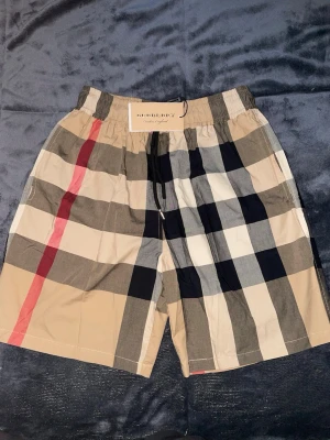 Burberry badshorts / swim shorts – Strl L/M - Säljer ett par snygga Burberry shorts med klassiska Nova Check-rutorna. Storlek L men passar även M beroende på hur man vill att de ska sitta. Elastisk midja med snörning för bra passform.  * Märke: Burberry * Storlek: L (passar även M) * Skick: Helt nya med tags kvar * Färg: Beige/rutig * Perfekta till sommaren, stranden eller vardagsbruk  Skriv vid frågor eller för fler bilder 📩 Skickas snabbt och säkert 📦