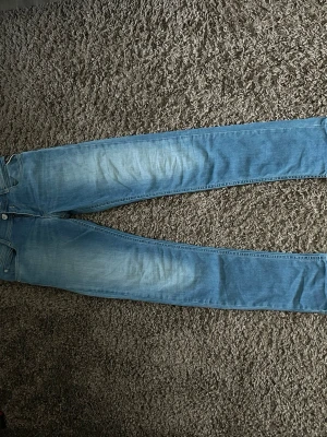 Ljusblå jeans från Replay - Säljer ett par ljusblå jeans från Replay med klassisk femficksmodell och diskret slitning på låren. Jeansen har smal passform och är tillverkade i stretchigt denimtyg för extra komfort. Perfekta för en avslappnad och trendig look.