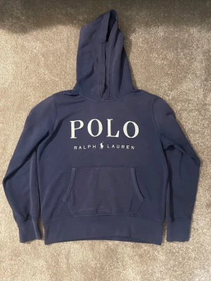 Mörkblå hoodie från Polo Ralph Lauren - Snygg mörkblå hoodie från Polo Ralph Lauren med stor logga framtill. Klassisk känguruficka och huva. Tillverkad i mjukt bomullsmaterial som är skönt att ha på sig. Perfekt för en avslappnad och trendig look.