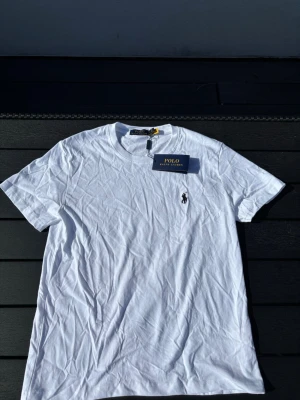 Vit t-shirt från Polo Ralph Lauren - Klassisk vit t-shirt från Polo Ralph Lauren med rund hals och korta ärmar. T-shirten har den ikoniska broderade loggan på bröstet och är tillverkad i mjuk bomull. Perfekt basplagg för en clean och stilren look.