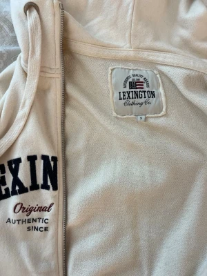 Vit hoodie från Lexington - Vit hoodie från Lexington med dragkedja, huva med dragsko och fickor framtill. Stora svarta bokstäver och broderad text på bröstet. Klassisk college-stil och mjukt material, perfekt för chill dagar eller när du vill ha en avslappnad look.