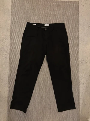 Svarta chinos från Jack & Jones - Snygga svarta chinos från Jack & Jones med klassisk rak passform och knappgylf. Tillverkade i mjukt bomullstyg som känns skönt mot huden. Perfekta för dig som vill ha en clean och stilren look.