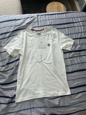 Vit t-shirt från Moncler - Säljer en stilren vit t-shirt från Moncler med klassisk rund hals och korta ärmar. T-shirten har Monclers ikoniska logga broderad på bröstet och är tillverkad i mjuk bomull. Perfekt till jeans eller shorts för en clean look.