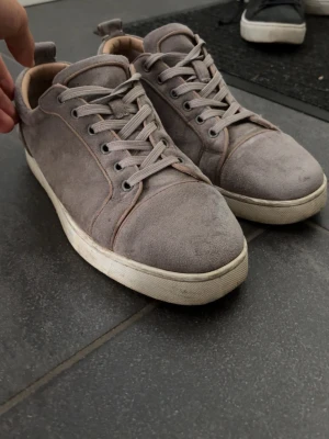 Gråa loubs  - Snygga gråa sneakers i mocka från Christian Louboutin med klassisk röd sula och vita kanter. Skorna har rund tå, matchande gråa skosnören och diskreta sömdetaljer. Perfekta för dig som gillar stilrena och exklusiva sneakers. Kommer med kvitto