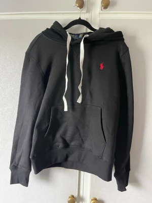Svart hoodie från Polo Ralph Lauren - Svart hoodie från Polo Ralph Lauren med vit dragsko i huvan och röd broderad logga på bröstet. Klassisk känguruficka framtill och ribbade muddar vid ärmar och nederkant. Perfekt för en avslappnad och stilren look.