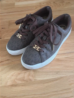 Bruna Michael Kors sneakers - Stilrena bruna sneakers från Michael Kors i storlek 37 med guldfärgade detaljer på snörningen samt logga både på in- & utsidan. Använt en gång, till salu pga för små. Nypris 1 300, säljs för 900.                                            Pris går att diskuteras ☺️
