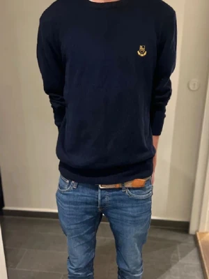 Mörkblå tröja Robbie Moor - Mörkblå stickad tröja från Robbie Moor med gul broderad logga på bröstet. Klassisk rund halsringning och ribbade muddar vid ärmslut och nederkant. Perfekt till jeans för en stilren look.