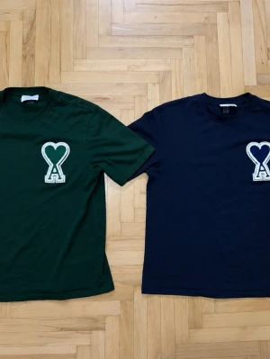 Två t-shirts från Ami Paris  - Två stilrena t-shirts i mörkgrönt och mörkblått med en stor vit hjärtformad logga med bokstaven A på bröstet. Perfekta för en trendig och avslappnad look. Använd några gånger men ändå i mycket bra skick. Försten till kvarn (fast pris)